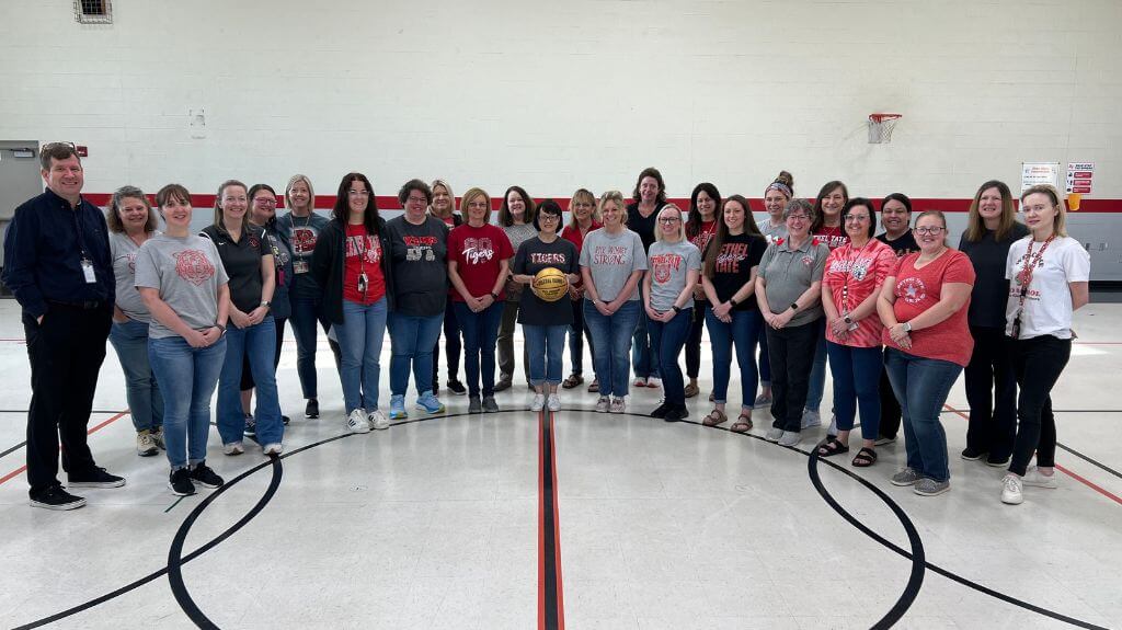 A group of educators gathers in a school gymnasium, standing in a semi-circle and smiling at the camera. Many are wearing red, gray, or black shirts with school spirit logos. A person in the center holds a golden basketball, symbolizing a team achievement or event. The gym features a basketball hoop and a red and white color scheme on the walls.